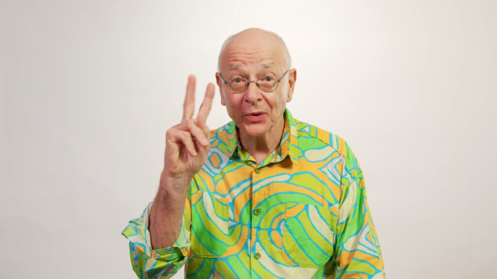 An older man with glasses and a bald head wears a colourful, patterned shirt in shades of green, yellow, and blue. He faces the camera, smiling, and holds up two fingers in a peace sign against a plain white background.