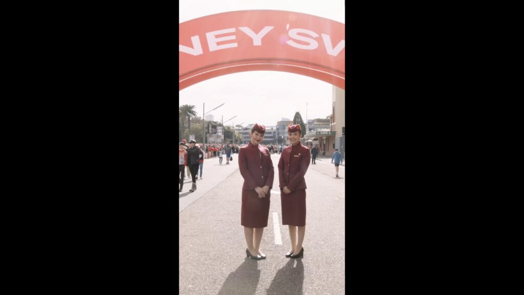 Two female flight attendants in matching burgundy uniforms and hats stand side by side under a large red archway on a city street. People walk in the background, and the street is closed to traffic for an event. The scene is bright and daytime.
