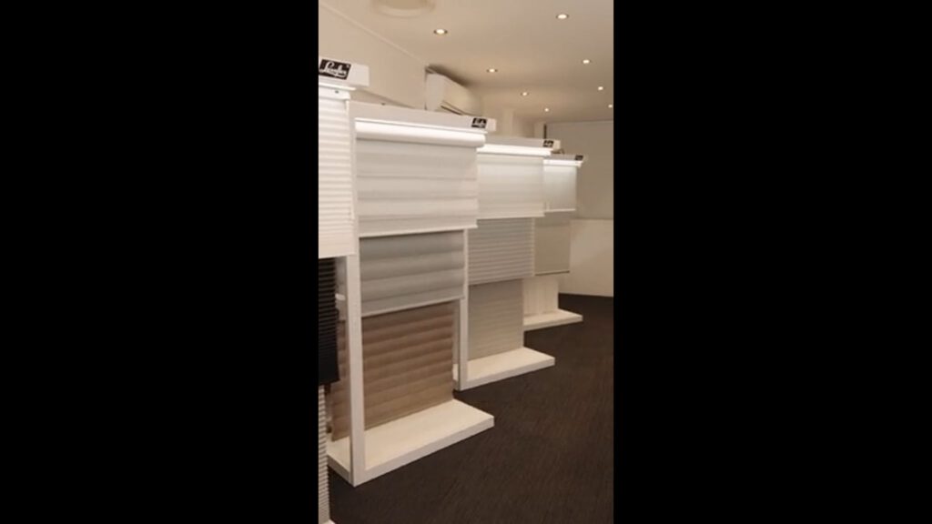 Several display stands show different types and colours of window blinds in a well-lit showroom with dark carpet flooring and white walls. Each stand features samples of blinds in shades of white, grey, and beige. Ceiling spotlights illuminate the space.