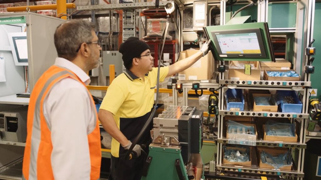 Two men in a factory, one in a yellow shirt and black beanie operating a touchscreen, the other in a white shirt and orange safety vest watching. Shelves with small parts and equipment surround them, with industrial machinery in the background.