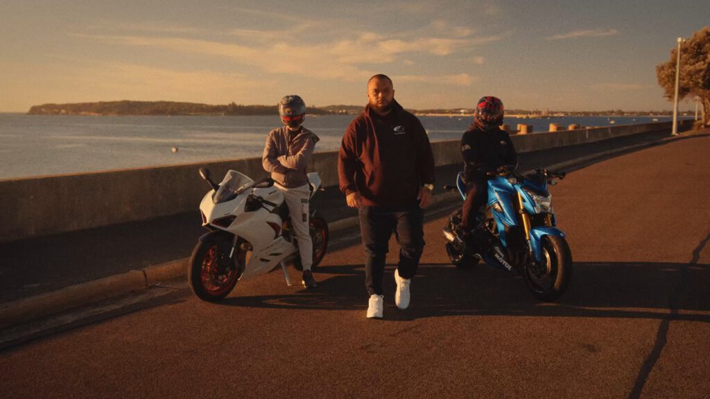 A man in a dark hoodie and white trainers stands on a waterfront road at sunset, with two people in helmets and riding gear beside him, each next to a sport motorbike—one white and one blue. The calm water and coastline are visible in the background.