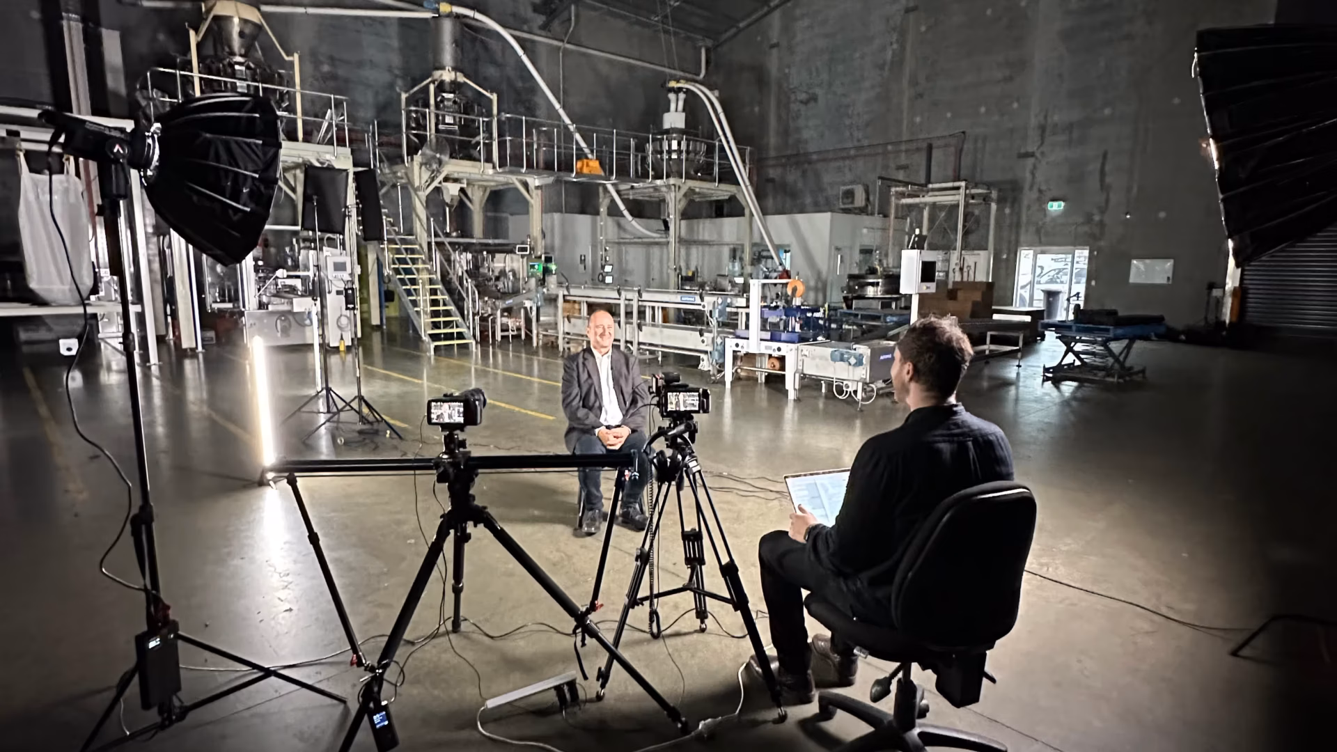 Two men sit facing each other in an industrial warehouse, filming an interview. Professional lights and cameras from a video production Sydney crew surround them. Metal stairs, industrial equipment, and high ceilings create dramatic shadows on the spacious floor.