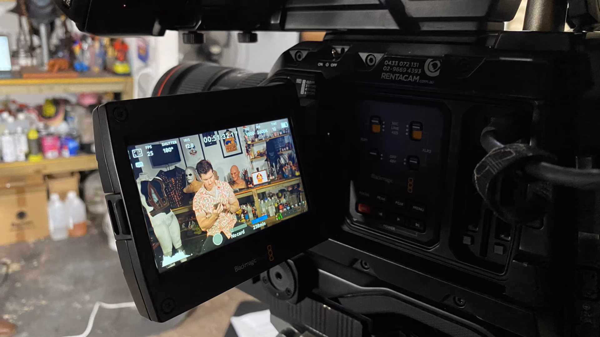Close-up of a professional video camera used for video production Sydney, its display screen showing two people working at a table with drinks and snacks in a colourful, decorated room. The background is slightly blurred, with the camera dominating the right side.