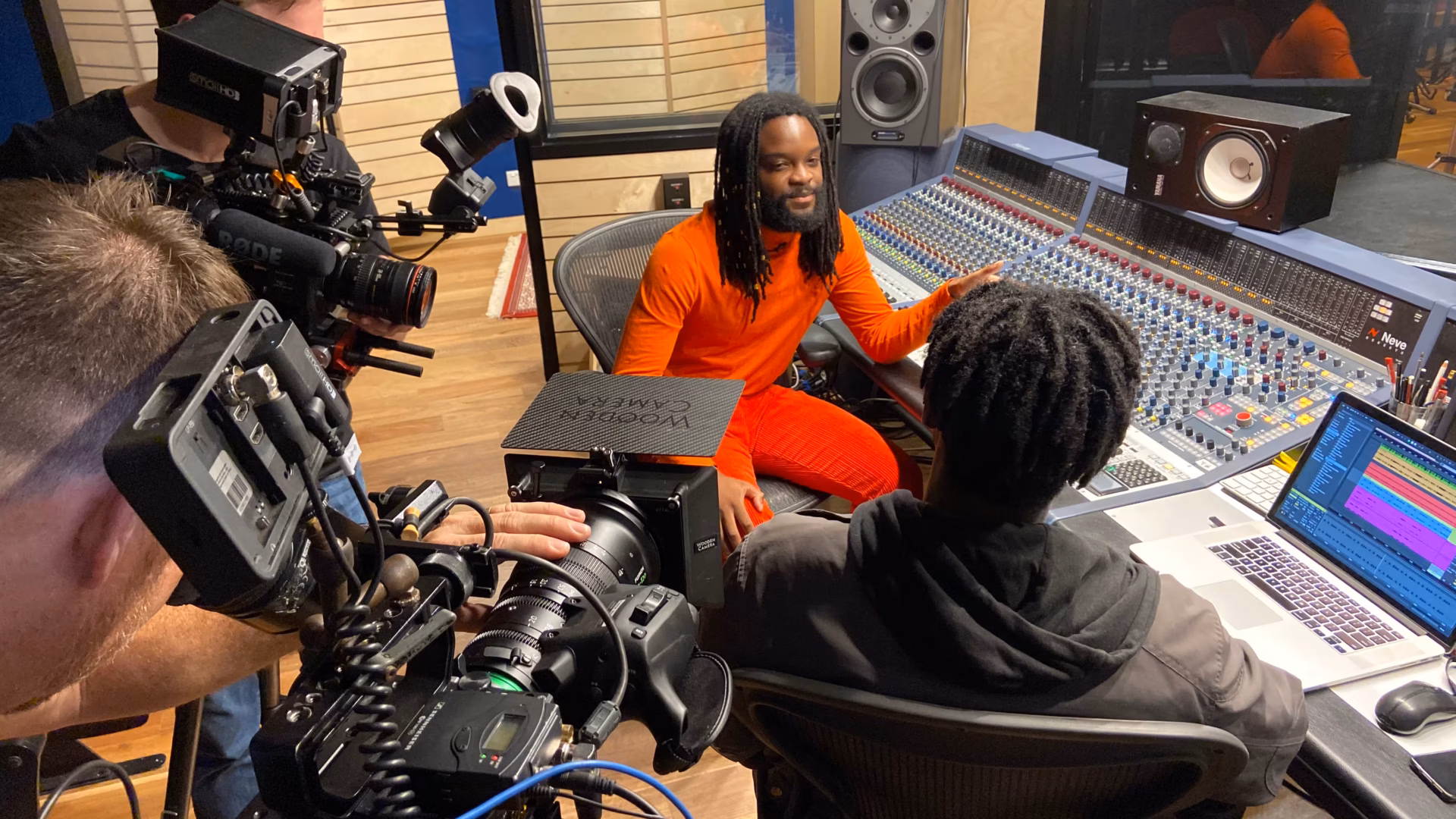 Two men sit at a large audio mixing desk in a recording studio, one in an orange tracksuit facing the camera crew. Two cameramen, part of a video production Sydney team, record the conversation and showcase the studio equipment.