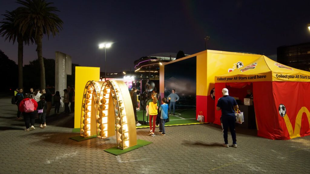 People gather at an outdoor football-themed event at night. There’s a lit arch, a yellow and red McDonald’s tent, and a display for collecting All Stars cards. Children and adults interact near a small fenced football area. Palm trees and city lights are visible.