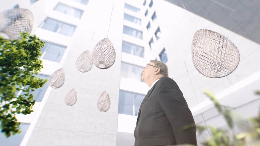 A man in a dark suit and glasses looks upwards in a modern, white, open atrium. Artistic, oval-shaped wire sculptures hang from above. Large windows and greenery are visible, and sunlight brightens the architectural space.