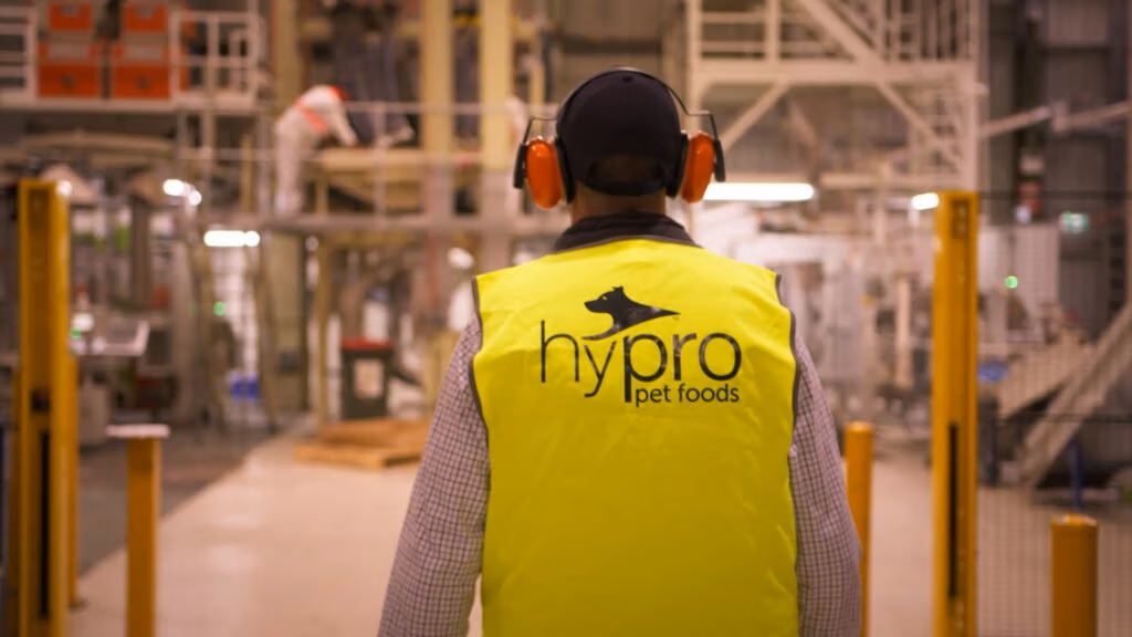 A person wearing a yellow safety vest labelled hypro pet foods and orange ear protection stands in a factory or industrial setting, facing away from the camera. Industrial equipment and structures are visible in the blurred background.