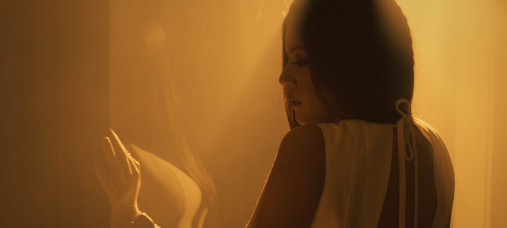 A woman in a white, backless dress stands in soft, golden light, her face in profile and eyes closed. She gently touches a translucent surface with her hand, casting a subtle reflection. The atmosphere is calm, dreamy, and intimate.
