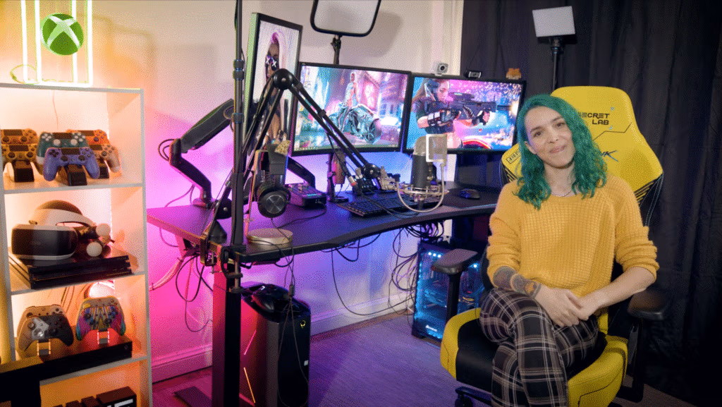 A woman with teal hair and a yellow jumper sits in a yellow gaming chair next to a desk with two monitors, a keyboard, microphones, and colourful LED lighting. Shelves on the left display game controllers and consoles. The room has a gaming and streaming setup.