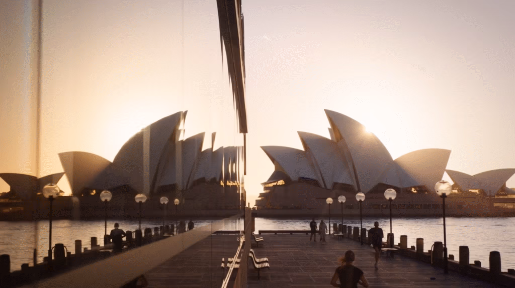 The Sydney Opera House is pictured at sunrise, with its white sail-like roofs illuminated by golden light. The building and the sun are reflected in a glass wall on the left, whilst people walk and stand along a waterfront promenade in the foreground.