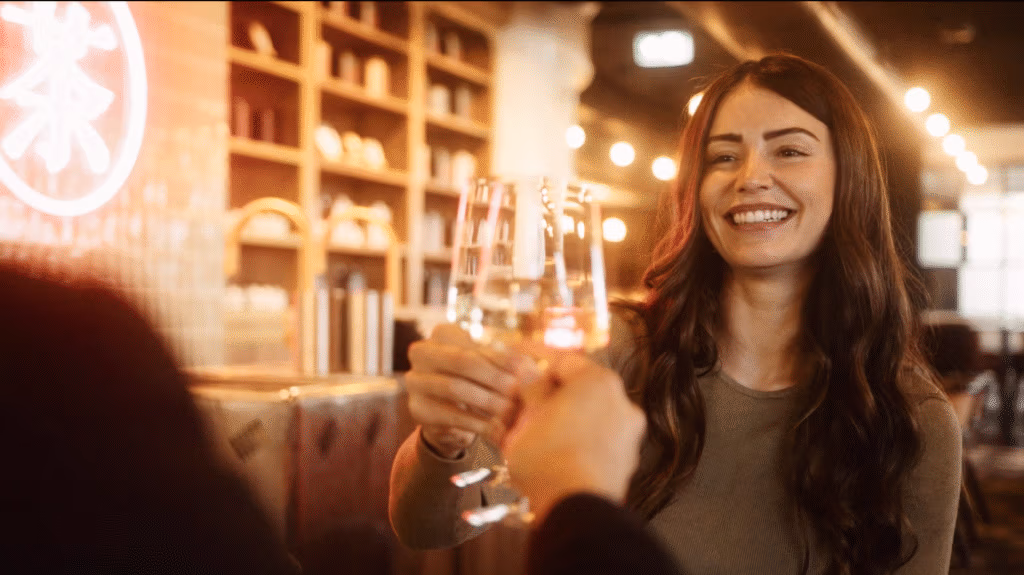 A woman with long brown hair smiles and clinks glasses with two others in a warm, softly lit bar. Wooden shelves and a neon sign are visible in the background, creating a cosy, inviting atmosphere.