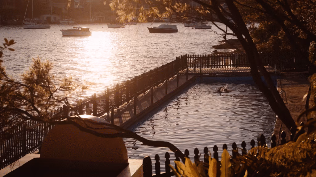 A fenced outdoor pool beside a river, with a person swimming. Sunlight glistens on the water. Trees frame the scene, and boats float on the river in the background, creating a peaceful, sunlit atmosphere. Shadows and warm tones suggest late afternoon or early evening.