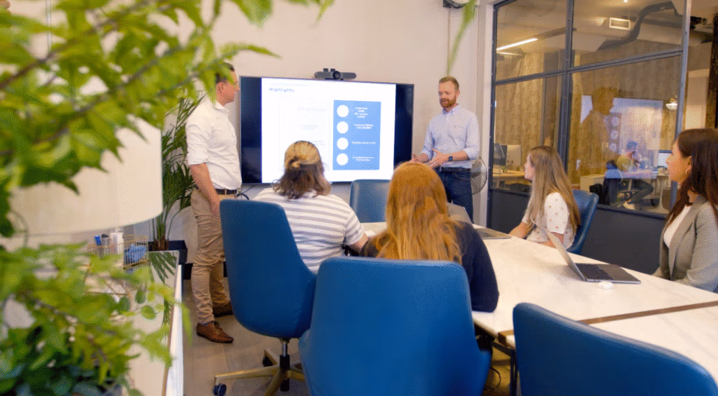 Five people are in a modern conference room. Two men stand near a large screen displaying a presentation, while three women sit at a table with laptops and notebooks. A green plant is visible in the foreground on the left side of the image.