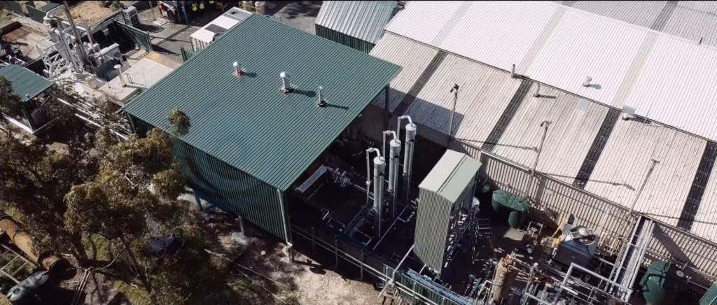Aerial view of an industrial facility with green and white metal roofs, vertical silver pipes, cylindrical tanks, and fenced perimeter, surrounded by some trees and machinery, under daylight.