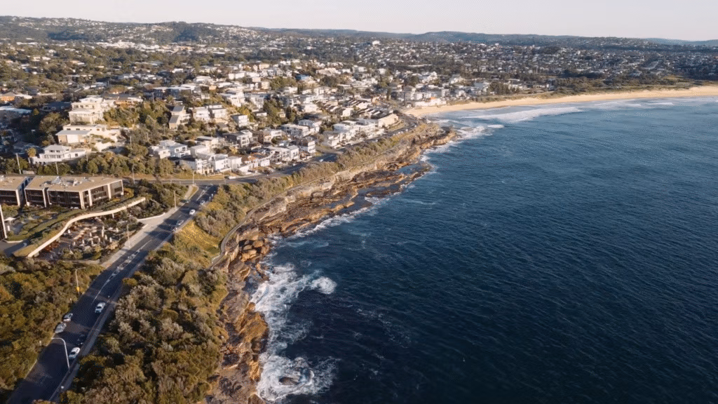 Aerial view of a coastline with deep blue ocean waves crashing against rocky cliffs. Residential houses and buildings line the shore, surrounded by greenery. A sandy beach curves along the right side, with a town visible in the background.