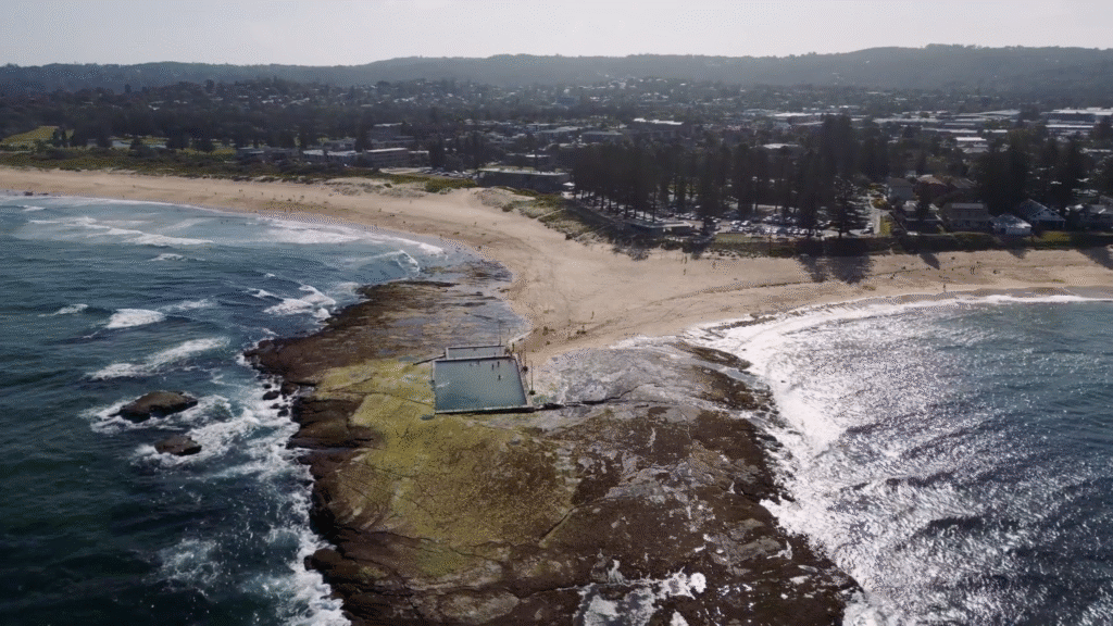 Aerial view of a coastal rock pool adjacent to the sea, with waves splashing over rocky shorelines. Sandy beach stretches inland, bordered by houses, trees, and distant hills under a hazy sky. People are visible around the pool and on the beach.