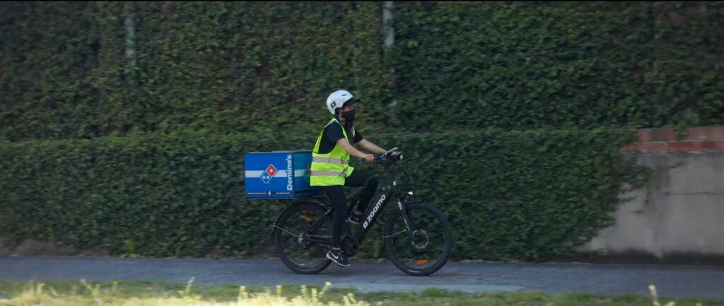 A Domino’s pizza delivery worker wearing a helmet and a yellow safety vest rides an electric bicycle on a path beside a tall green hedge. The blue insulated Domino’s box is mounted on the rear of the bike.