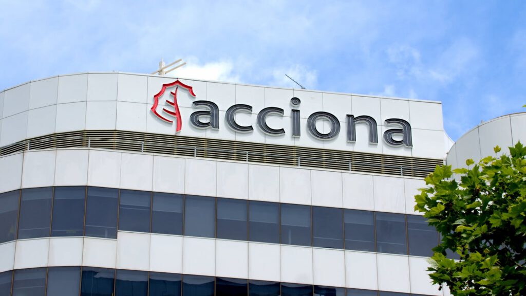 A modern office building with white and glass exterior features the large “acciona” logo on top. The logo includes a red outline of a tree. The sky is blue with some clouds, and part of a leafy green tree is visible in the lower right corner.