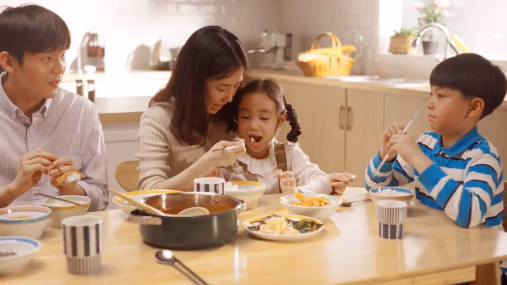 A family of four sits at a kitchen table eating a meal. The mother in a cream jumper feeds her young daughter with a spoon, while a man and a boy in a blue striped shirt eat their own food. Sunlight brightens the cosy, inviting kitchen in the background.