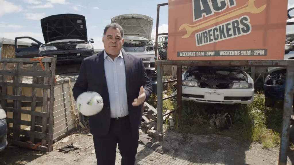 A man in a suit stands outdoors holding a white football, with several cars behind him that have their bonnets open. He is next to a red sign reading “ACT WRECKERS.” The scene appears to be a scrapyard under a partly cloudy sky.