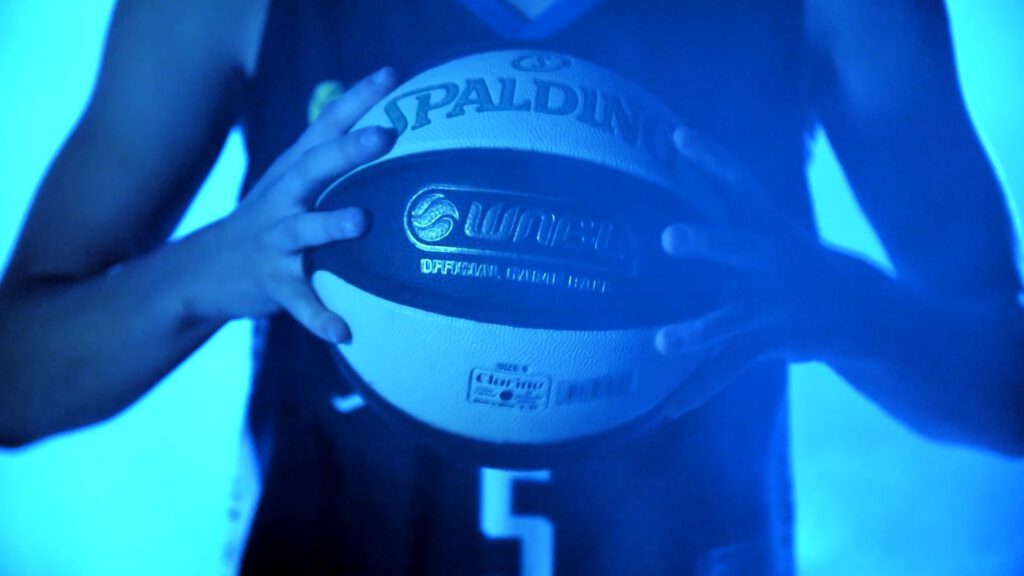 A person in a dark basketball vest with the number 5 holds a Spalding basketball with both hands. The image is bathed in blue light, creating a dramatic effect and partially obscuring facial and vest details.