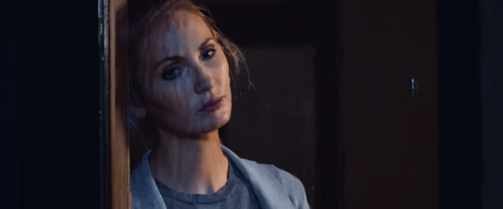 A woman with fair skin and blonde hair leans her head against a wooden doorframe, gazing forward with a sombre expression. She wears a grey shirt and a pale blue jacket. Soft, moody lighting casts shadows on her face and the dark background.