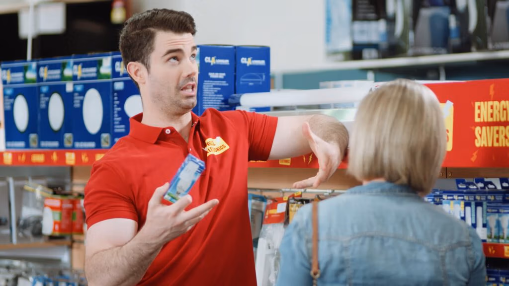 A man in a red polo shirt gestures animatedly whilst holding a light bulb package, speaking to a customer with short blonde hair in a denim jacket, inside a shop aisle with shelves of lighting products.