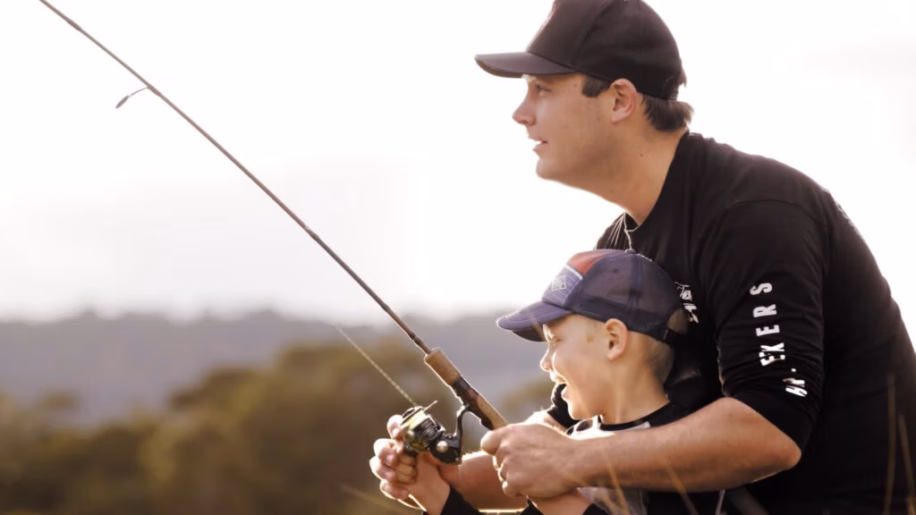 An adult and a child, both wearing caps, smile as they hold a fishing rod together outdoors. The adult helps the child reel in the line. The background shows blurred trees and soft sunlight, creating a warm, joyful atmosphere.