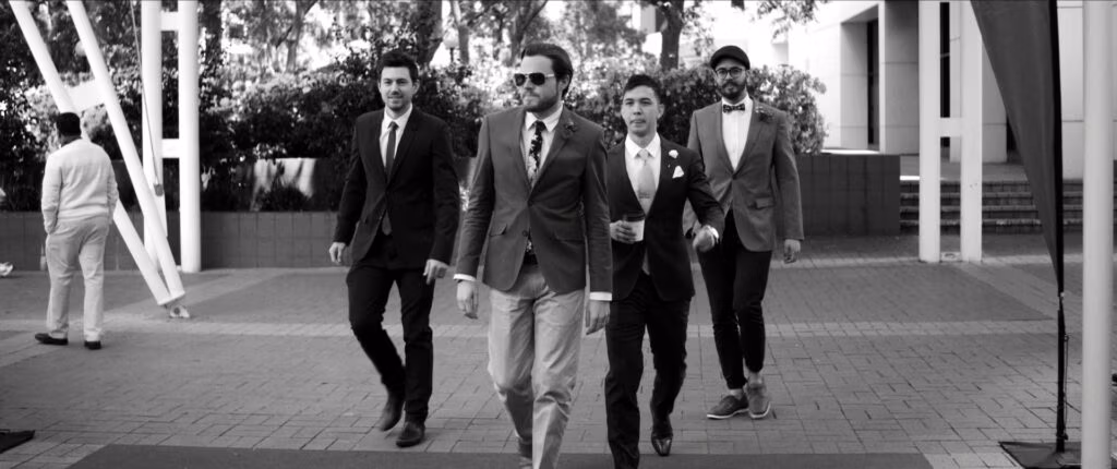 Four men in suits stride confidently towards the camera outdoors. The central man wears sunglasses and a floral tie, flanked by men in suits with ties or bow ties. Trees and modern buildings are visible in the background on a sunny day. Image is in black and white.