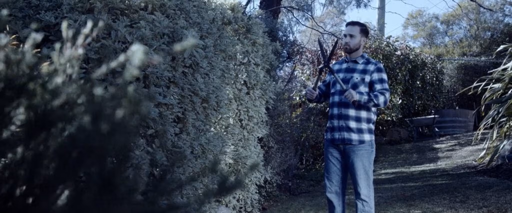 A man in a blue checked shirt and jeans stands in a back garden, holding open secateurs, facing a large, dense hedge. Sunlight filters through trees, casting shadows on the grass and plants around him. The atmosphere feels calm, and he appears focused on trimming.