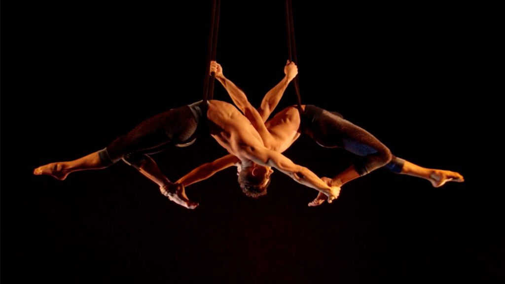 Two acrobats perform an aerial act, suspended upside down by straps. Their bodies are intertwined, arms and legs extended outward in a symmetrical, star-like formation. The background is dark, highlighting their muscular forms and the dramatic pose.