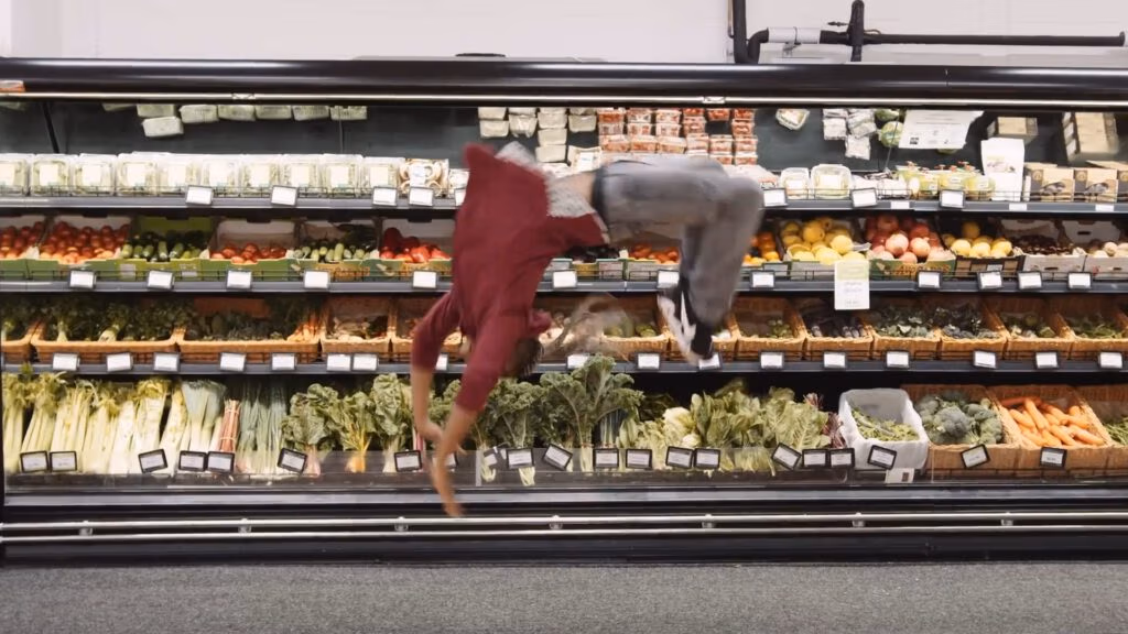 Two people are performing acrobatic flips in front of a supermarket produce section filled with leafy greens, vegetables, and fruit, with neatly arranged labels and shelves in the background.