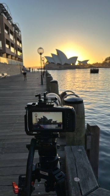 A camera on a tripod is set up on a wooden walkway by the water, capturing the Sydney Opera House at sunrise. The Opera House is silhouetted against the bright sky, and buildings line the left side of the walkway.