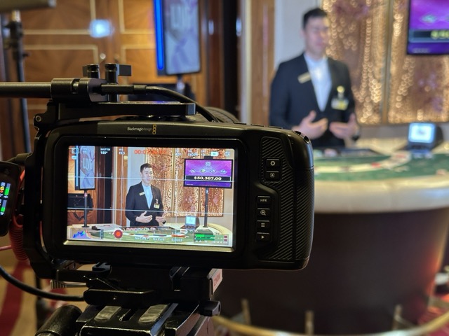 Close-up of a video camera filming a casino croupier standing behind a gaming table. The camera’s screen displays the croupier in focus, with casino elements and digital graphics visible. The background shows a casino setting with warm lighting and ornate decor.