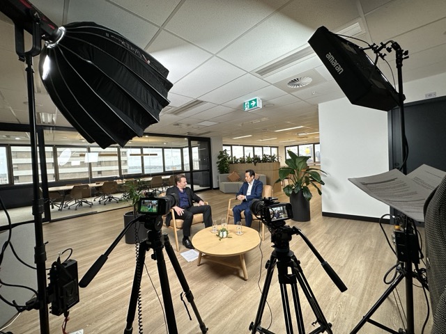 Two people sit on chairs at a round wooden table in a modern office, engaged in conversation. Studio lights, cameras, and a microphone are set up around them, suggesting a recorded interview. Large windows and indoor plants are visible in the background.