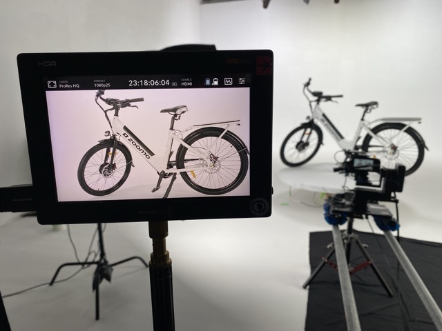 A camera monitor displays a close-up shot of a white electric bike in a photography studio, while two similar bikes are positioned on a white backdrop in the background. Lighting equipment and a slider are visible, indicating a professional photoshoot setup.