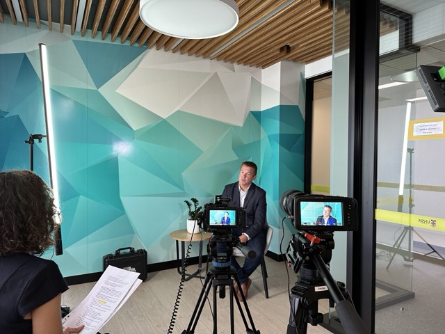 A man in a suit sits on a stool in a modern office with geometric blue-green walls, being filmed by two cameras. Light stands and interview notes are visible. A woman holding papers sits off-camera facing him, conducting the interview.