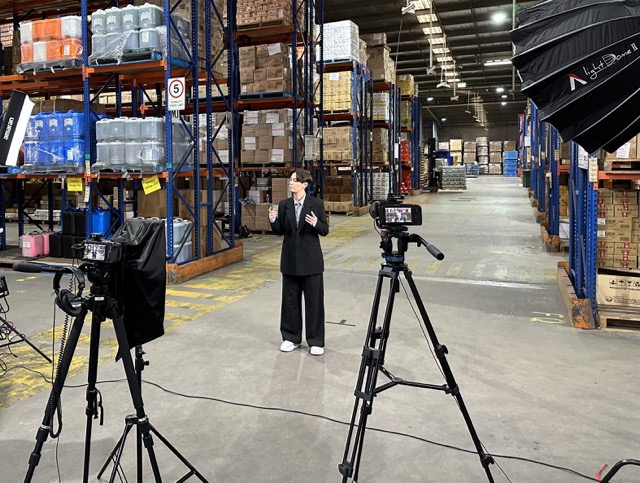 A person in a black suit stands in a warehouse surrounded by shelves of boxes and goods. Professional video cameras and large studio lights are set up, filming the person, who is speaking or gesturing with hands. The warehouse aisles extend into the background.