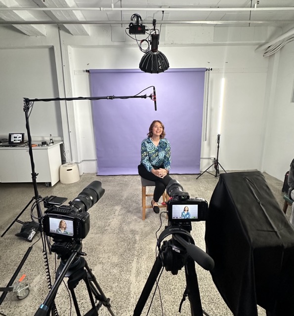 A woman in a patterned green and white top sits on a stool, smiling, in front of a lavender backdrop. Multiple cameras, lights, and a microphone surround her in a studio set-up for a video or photo shoot.