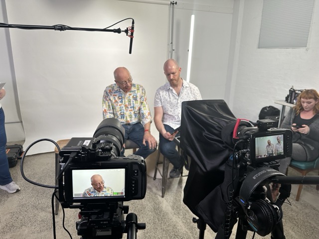 Two men sit side by side in front of a white backdrop in a studio, filmed by two cameras. A microphone hangs above them. A woman sits in the background on the right, looking at her mobile. Studio lights and equipment are visible around them.