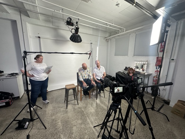 A behind-the-scenes view of a studio setup: two men sit on stools facing cameras, while a woman stands nearby holding papers. Studio lights, tripods, and equipment are visible, with a white backdrop and production staff preparing for filming.