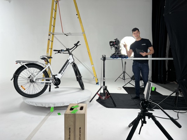 A man stands in a photo studio beside a camera and lighting equipment, gesturing to a white electric bike on a circular platform. Behind the bike is a yellow ladder. The scene is well-lit, with a white backdrop and various tripods and gear visible.