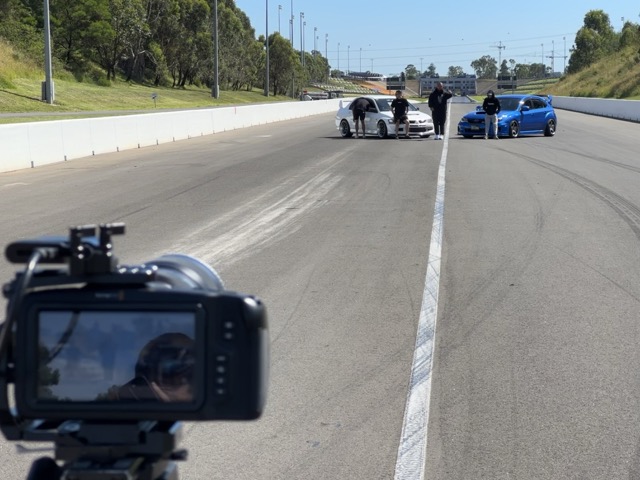 A video camera is set up in the foreground, focused on two cars—a white car and a blue car—parked side by side on a racetrack. Three people stand near the cars under a clear sky, with trees and fencing in the background.