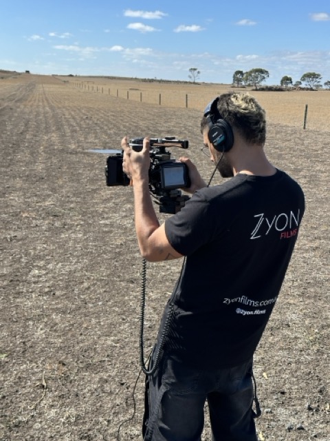 A person wearing headphones and a black Zyon Films T-shirt operates a professional video camera in a dry, open field under a blue sky. The landscape is flat and barren, with fences and sparse trees in the distance.