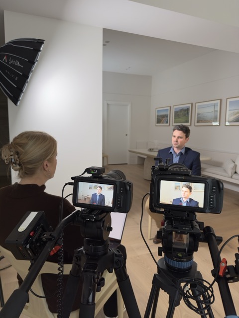 A man in a suit is being interviewed by a woman with blonde hair, seated opposite him. Two cameras and a softbox light are set up, filming the scene in a bright room with light wood floors and framed photos on the wall in the background.