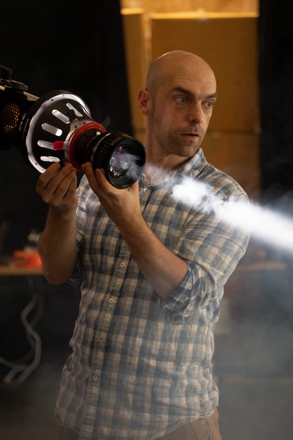 A man in a checked shirt holds a large spotlight emitting a visible beam of light through misty air. He looks intently to his left, standing in a dimly lit room with blurred boxes and cables in the background.