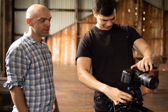 Two men indoors, one in a chequered shirt observing while the other, in a black T-shirt, adjusts a professional camera mounted on a tripod. The background is a rustic, wooden interior with soft lighting.