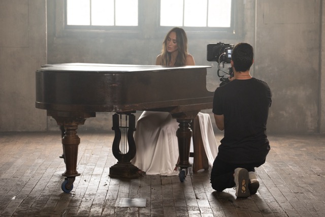 A woman in a long white dress sits at a grand piano in a dimly lit, rustic room with wooden floors. She faces a window as a cameraman kneels in front of her, filming. Light streams through the window, creating a moody atmosphere.