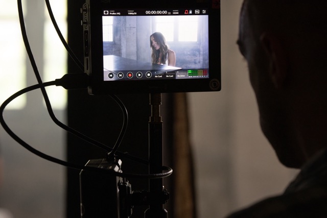 A monitor displays a woman with long hair sitting at a piano, filmed in a softly lit room with large windows. Cables connect to the monitor, and the silhouette of a person's head is visible in the foreground, observing the screen.