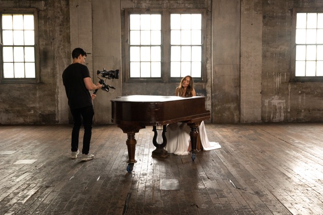A woman in a long white dress sits at a grand piano in a spacious, worn room with three large windows. A man in black holds a camera on a stabiliser, filming her. Light streams in, highlighting the rustic wooden floor and weathered walls.